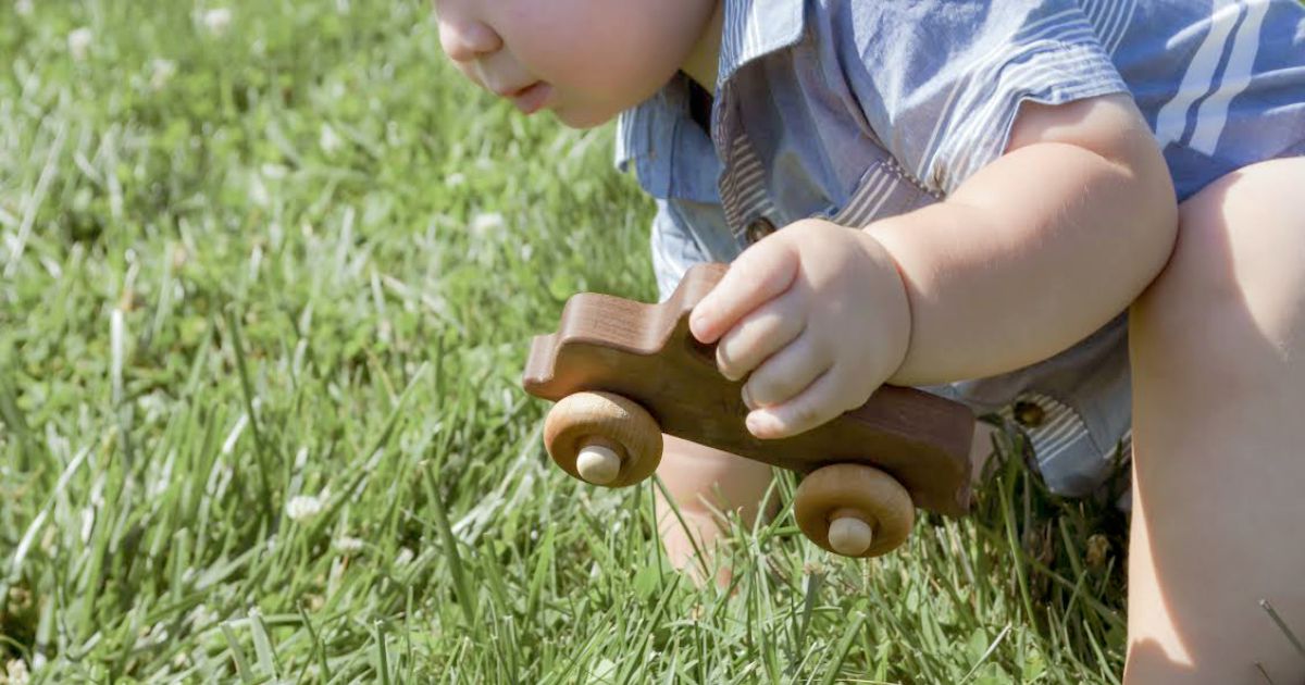 Close-up of a toddler holding a Smiling Tree wooden toy car while exploring grass—pure unplugged play at its best. A beautiful reminder of the joys and developmental benefits of nature-based play and outdoor learning for young children.