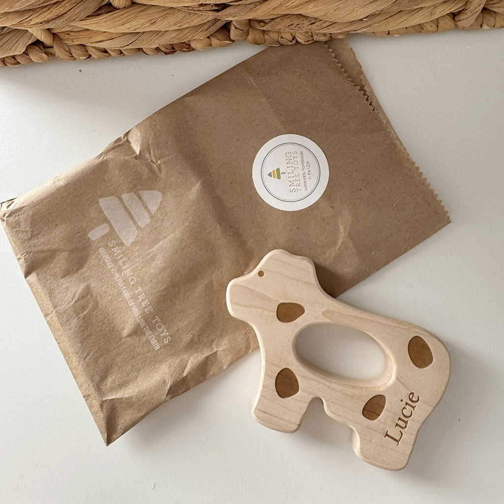 Wooden cow shaped teething toy with &#39;Lucie&#39; engraving on a brown paper bag.
